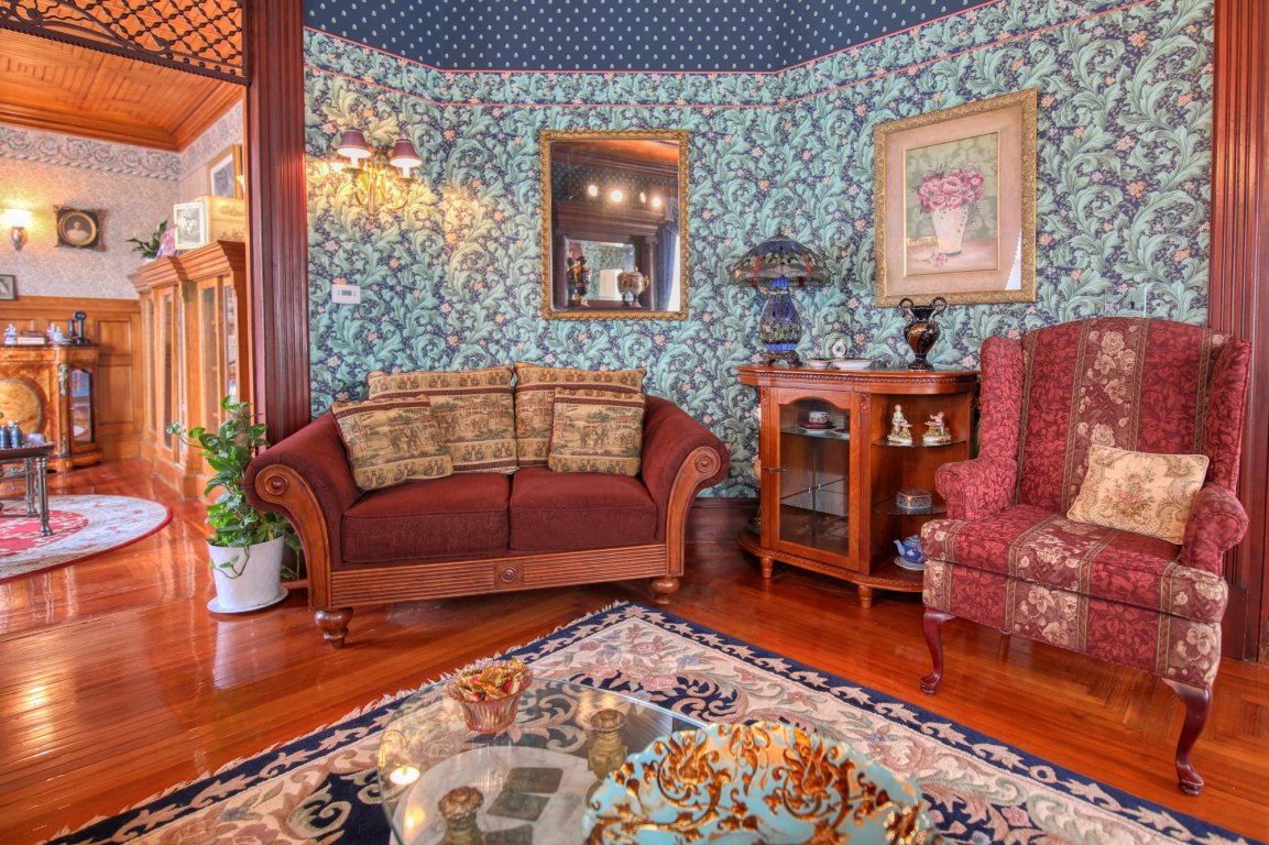 Victorian-style living room with patterned wallpaper, a red sofa and armchair, ornate rug, wooden floors, and vintage decor including a glass cabinet, lamp, framed mirror, and a large floral painting—perfect for those seeking the timeless charm often found in bed and breakfast rooms Toms River NJ.