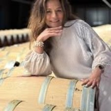 A woman with long hair and a light blouse leans on large wooden barrels, smiling at the camera in what appears to be a winery or cellar setting—evoking the warm, welcoming charm of an innkeeper at Mathis House in Toms River, NJ.