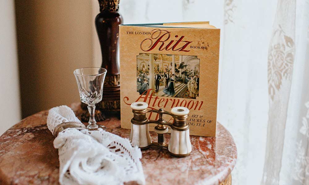 A book titled “The London Ritz Afternoon,” a crystal glass, lace napkin, and vintage opera glasses sit on a marble-topped table near a window with lace curtains—capturing the timeless elegance you might find in one of the exclusive bed and breakfast packages Toms River NJ has to offer.