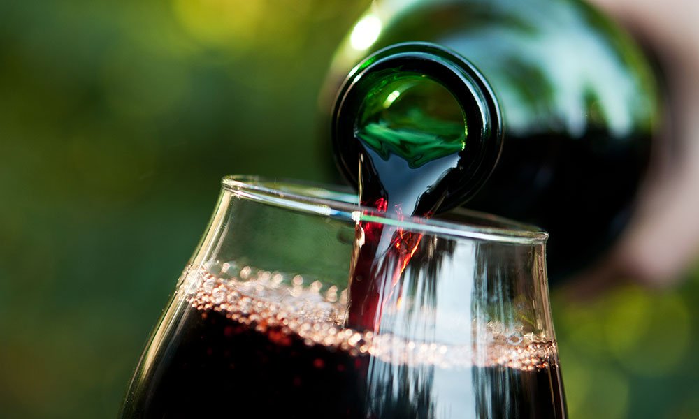 A close-up of red wine being poured from a green bottle into a glass, with bubbles forming on the surface and a blurred green background—an inviting scene reminiscent of leisurely afternoons exploring things to do in Ocean County NJ.