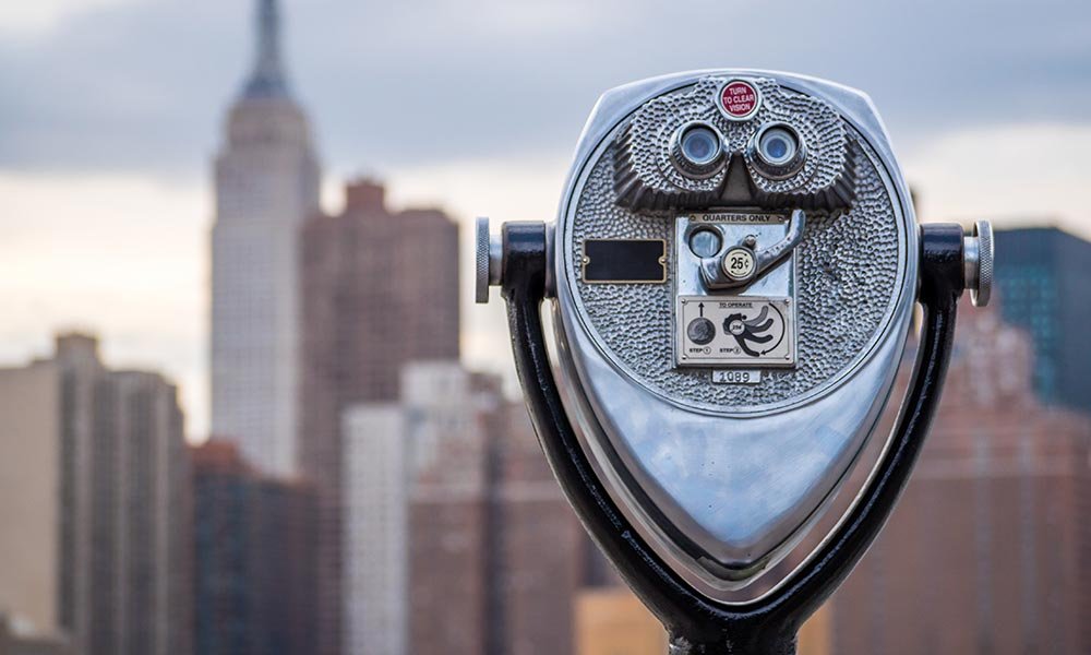 A coin-operated binocular viewer is in focus, with an out-of-focus city skyline, including the Empire State Building, in the background under a cloudy sky—a scene that might inspire you to seek out new adventures and things to do in Ocean County NJ.