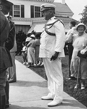 A man in a white military uniform stands on a sidewalk, with people and a house—possibly a charming bed and breakfast in Toms River NJ—in the background. Some people are dressed in summer clothes and hats.