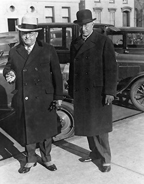 Two men in long dark coats and hats stand in front of a vintage car on a city street, reminiscent of classic scenes you might find near a historic bed and breakfast in Toms River NJ. One man holds a cigar, and both look toward the camera. The background shows buildings and more parked cars.