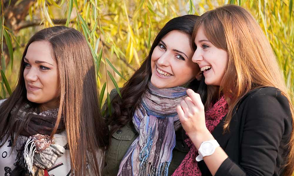 Three young women standing outdoors, smiling and laughing together, with green foliage in the background. They are wearing scarves and appear to be enjoying a pleasant moment, perhaps while discussing their favorite bed and breakfast packages in Toms River, NJ.