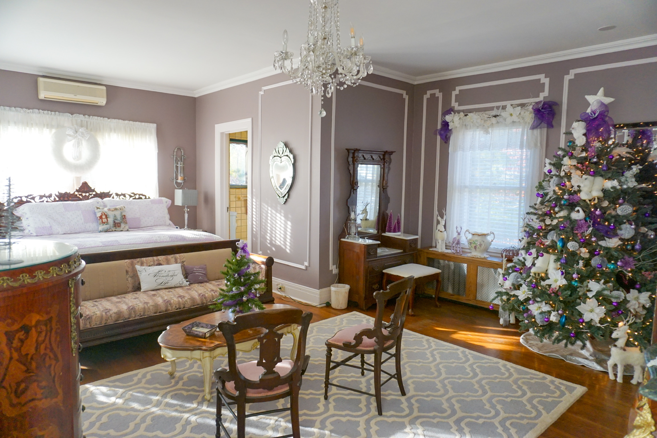 Elegant bedroom in the Queen Alexa suite, Toms River NJ, with a chandelier, lilac walls, ornate mirror, antique furniture, and decorated Christmas trees. Sunlight streams through sheer curtains onto a patterned rug and wooden floor.