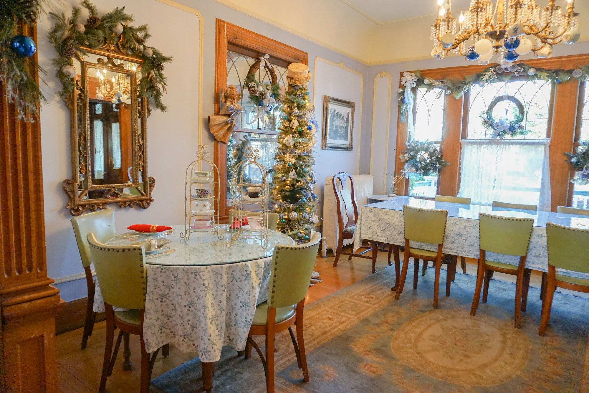 A bright, elegantly decorated dining room inside a charming bed and breakfast in Toms River NJ features holiday garlands, a Christmas tree, chandeliers, round and rectangular tables, and green chairs. Light pours in through stained glass windows.