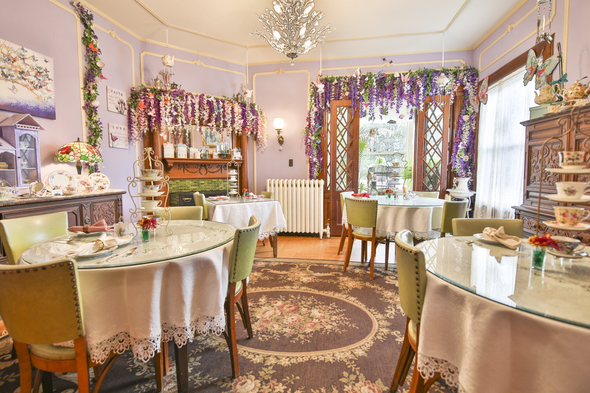 A cozy tea room with vintage decor, round tables with lace cloths, green chairs, floral arrangements, and hanging purple flowers awaits you inside this charming bed and breakfast in Toms River NJ. Sunlight streams in through windows and a glass door, creating a warm and inviting atmosphere.