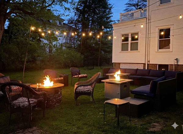 Outdoor patio at dusk with string lights above, several wicker chairs, two fire pits with flames, a long blue sofa, and a small white house in the background—surrounded by trees and grass—offers a tranquil retreat for guests enjoying their stay in bed and breakfast rooms Toms River NJ.