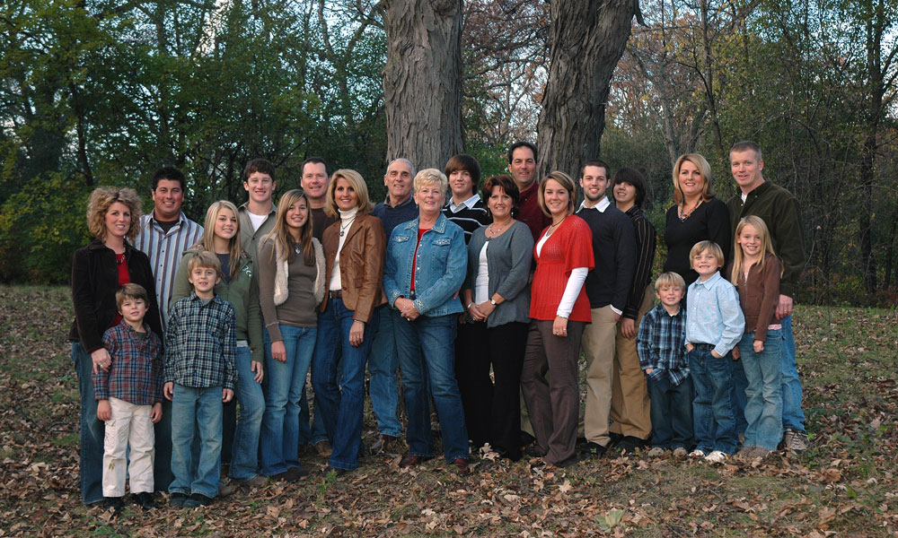 A large group of adults and children pose together outdoors in front of trees. They are standing on fallen leaves, dressed in casual autumn clothing, and smiling at the camera—capturing a perfect moment during their getaway with bed and breakfast packages in Toms River NJ.